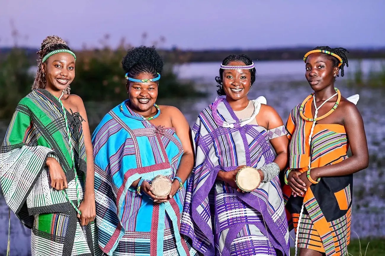 Women in colourful Venda minwenda stand by the water at dusk, holding drums.